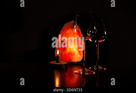 Two elegant empty clear wineglasses for red wine on the table with burning candle on dark black background with blur orange salt lamp. Closeup decor c Stock Photo