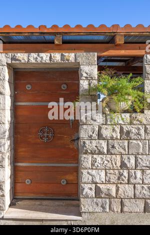 A rustic wooden door set in a textured stone wall, featuring decorative iron hardware. A leafy plant grows in a stone planter nearby, under a bright b Stock Photo