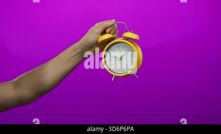 Man holding yellow clock against a vibrant purple wall, with arm pointing, creating a focused and colorful isolated composition. Stock Photo