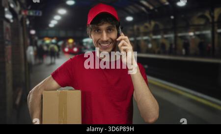 Man holds cardboard box with one arm and talks on phone at train station; efficiency reliability service. Stock Photo