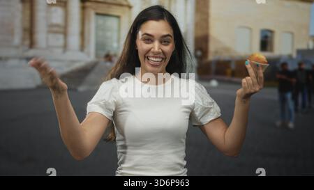 Young hispanic woman beckons with hand while holding muffin on street near historic building, smiling to camera; joy friendly invite. Stock Photo