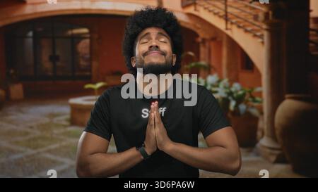 African american man in staff uniform standing outdoors with hands together appearing thankful in a courtyard with warm tones and potted plants in the Stock Photo