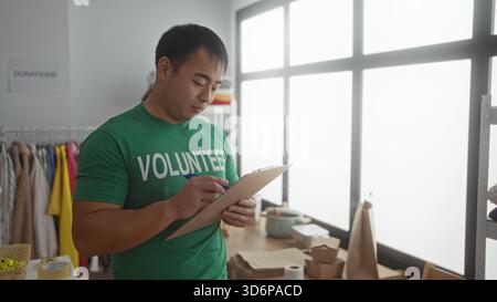 Asian man volunteering in a charity center, wearing a green shirt, taking notes on a clipboard in a room full of donations. Stock Photo