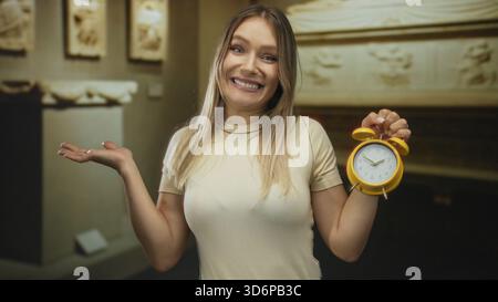 Blonde woman holding an alarm clock smiles in indoor museum setting. Stock Photo