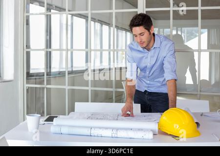 Man wearing blue shirt leaning over office table reviewing plans with yellow hard hat, copy space. Architect, blueprints, studio, daylight, glass, mod Stock Photo
