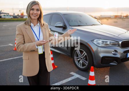 Female Driving Instructor Presenting Training Car at Driving School Area Stock Photo