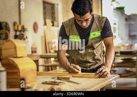 Carpenter with glasses taking measurements at the workbench. Young cabinetmaker measuring a piece with a tape measure in the workshop Stock Photo