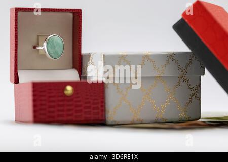 an elegant silver ring with a large aquamarine/emerald stone in an open red gift box, next to a decorative white and gold patterned box and a black bo Stock Photo