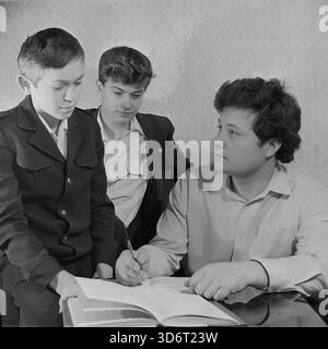 Slaviansk, Donetsk region, Ukrainian SSR, Soviet Union. February 1986. A railway worker deputy helping his two sons with homework at home. The father, dressed in a casual shirt, sits at a polished table holding a pen and writing in a notebook. He talks to his younger son, who wears a dark school uniform jacket. The older brother watches from behind. This archival black and white photograph captures a moment of parental guidance and education in the peaceful Donbass region long before the war. Stock Photo