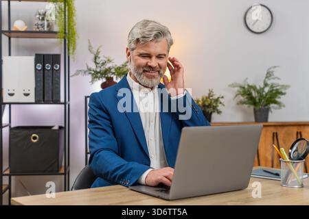 Middle-aged businessman at home office codes on laptop wearing earphones listening to music while focusing on project. Freelancer guy at table enjoys it rhythm typing code delivering software feature. Stock Photo