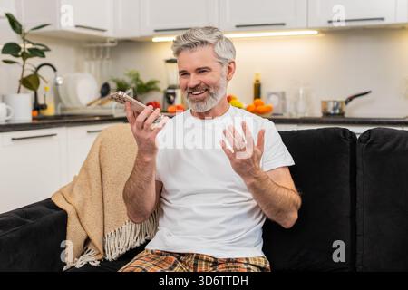 Middle-aged man at home records voice note on smartphone waves hello on speaker and shares news. Mature guy on sofa smiles talks sends chat reply then listens to playback nods and adds a short update. Stock Photo
