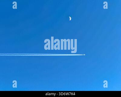 Holiday jet in flight with contrails passing the moon on a deep blue sky Stock Photo