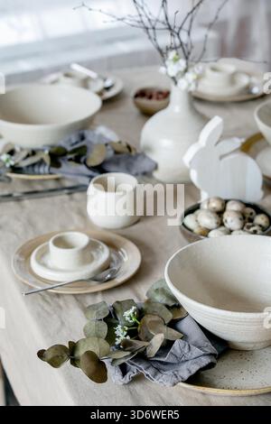 Rustic Easter table setting with empty craft ceramic tableware, plates and bowls, Easter bunny rabbit decorations, branches in vase on linen tablecloth in front of the window. Easter holiday dinner. Stock Photo