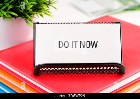 A white card with the text DO IT NOW stands on a stack of colorful notebooks next to a small potted green plant, presenting a call to action. Stock Photo