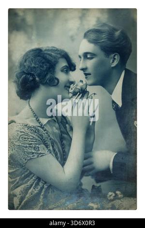 A vintage, blue-tinted postcard portrays a young couple, with the woman holding a rose and smiling at the man, suggesting a sweet, romantic interactio Stock Photo