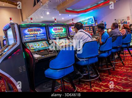 People seated at Multi Win slot machines inside a colorful casino with bright lights, blue chairs, and a red patterned carpet. Stock Photo