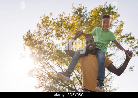 Elementary age boy having fun with his father outdoors, sitting on his dad shoulders while enjoying a walk along a field trail during spring break. Stock Photo
