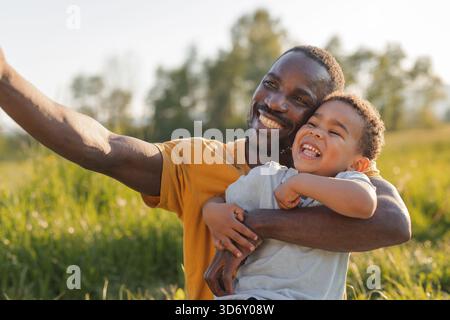A father smiles joyfully while holding his young son in a grassy field during daylight, capturing a special moment selfie. Stock Photo