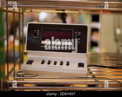 A vintage white electronic console with a screen and push buttons displayed on a metal shelf. The device showcases classic industrial technology Stock Photo