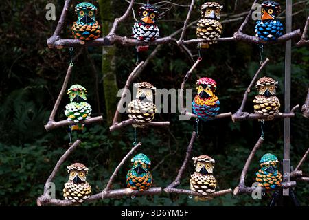Colorful handmade funny owl figures crafted from pine cones hang on wooden branches in a forest setting, creating a vibrant and playful outdoor Stock Photo