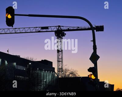 A construction crane towers in the foreground as the sun sets in Neuss, Germany. Traffic lights glow softly, marking the end of the day in this vibran Stock Photo