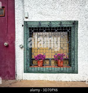Decorative window with metal grill and potted purple flowers on a stucco wall with contrasting red and white colors Stock Photo