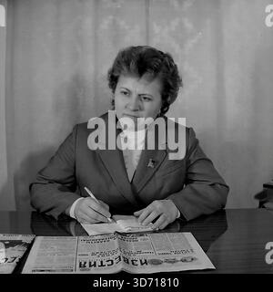 Slaviansk, Donetsk region, Ukrainian SSR, Soviet Union. February 1986. A portrait of Deputy Marchenko pausing while working at a desk. The woman, wearing a formal suit with a deputy badge, holds a pen over a notebook and looks aside with a serious, contemplative expression. A copy of the Soviet newspaper "Izvestia" lies open on the polished table in the foreground. This archival black and white photograph captures the responsibility and administrative work of a local leader in the peaceful Donbass region long before the war. Stock Photo