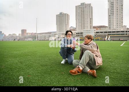 Two Russian women, friends of 50 and 60 years old, in tracksuits, sit tired on the grass after sports and drink water from a bottle and talking Stock Photo