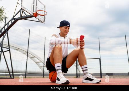 Female athlete sitting on basketball using smartphone on street court Stock Photo