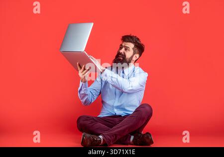 Happy young man sitting on the floor with and using laptop computer on red background. Young businessman using his laptop, pc. Holding laptop computer Stock Photo