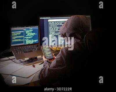 A person in a hoodie sits in front of two monitors and a laptop, displaying lines of code and a terminal. He holds a smartphone with the code displaye Stock Photo