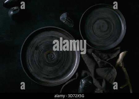 Set of empty black ceramic plates with black stones and wooden around on textile napkin over black wooden background. Flat lay. Stock Photo