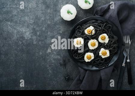 Halloween party Italian black pasta decorated horror olives like eyes on black plate on old dark table background. Monster face from pasta. Halloween decorations and Halloween food concept. Mock up. Stock Photo
