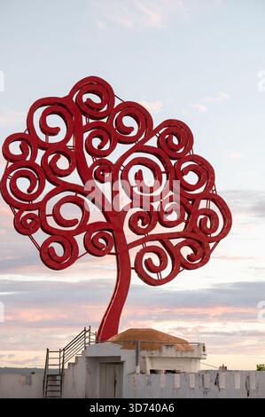 A red tree sculpture with swirling branches is silhouetted against a colorful sunset, showcasing artistic design. Stock Photo