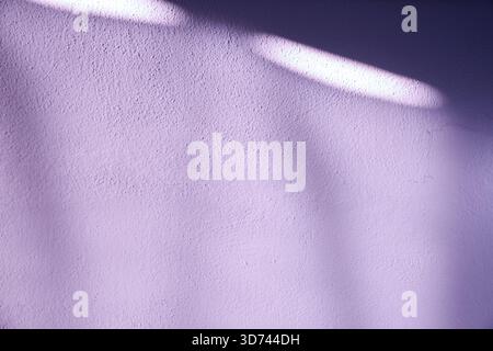 A lavender-colored textured wall illuminated by natural sunlight, with soft shadows forming abstract patterns across the surface. The pastel tone crea Stock Photo