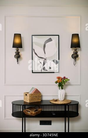 Modern console table standing against light wall with abstract framed artwork, two wall sconces, woven basket with books, ceramic vase holding fresh flowers, remote control on tabletop Stock Photo