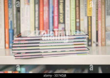 Stack of colorful magazines, brochures on shelf with blurred books in background. Concept of reading materials, publishing industry, educational Stock Photo