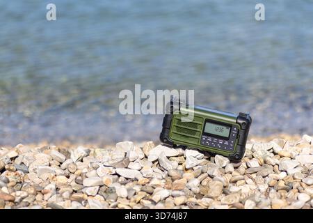 A durable green radio sits among smooth beach stones with gentle waves in the background, evoking outdoor adventure, survival, and preparedness for co Stock Photo