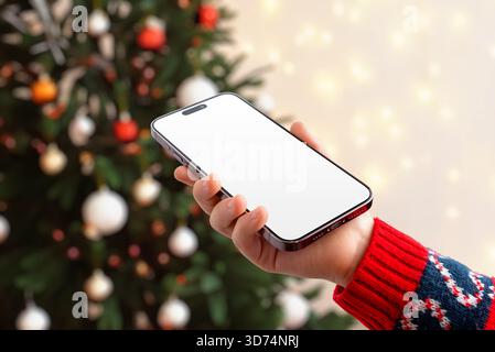 Girl using smartphone with blank screen in front of Christmas tree, ideal for app mockups, festive campaigns, mobile UI showcases and holiday marketin Stock Photo