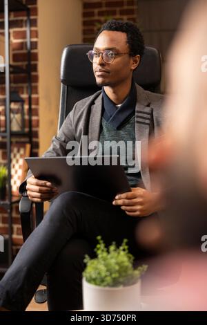 Psychiatrist sits in private clinic with laptop, listening and providing support to clients. Therapist wearing glasses conducts counseling session, helping patients cope with mental health challenges. Stock Photo