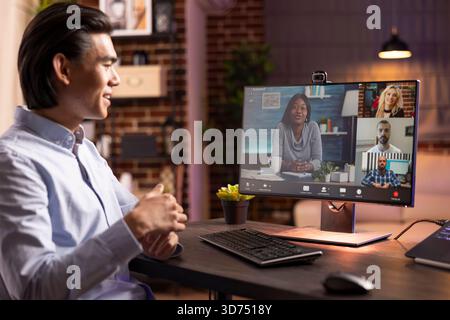 Portrait of young asian entrepreneur seated at desk during online meeting. Male remote worker with cheerful expression, collaborates with colleagues on digital business projects from home office. Stock Photo