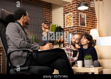 Caucasian couple seeking guidance from african american male counselor. Psychiatrist takes notes as wife discusses marriage issues while seated closely with husband during counseling session. Stock Photo