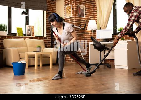 Multiethnic couple enjoys playful spring cleaning in brick wall apartment. Girlfriend with headphones, sings into mop stick as smiling boyfriend vacuums floor, turning chores into fun bonding moments. Stock Photo