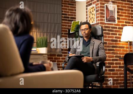 African American counselor wearing glasses listens attentively to young couple. Professional counseling session supporting patients with depression, stress and emotional challenges. Stock Photo