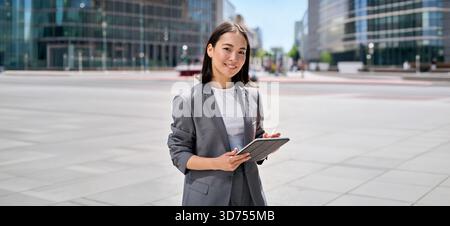 Young happy Asian business woman standing on city street using digital tablet Stock Photo