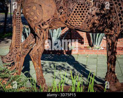 Rusted scrap‑metal horse sculpture, all gears and perforated plates, casting a sharp shadow by agaves and a brick planter. Stock Photo
