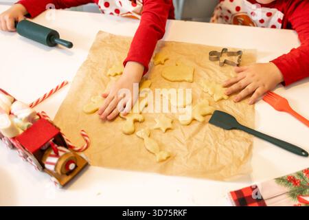 Two children in festive outfits are working together to shape cookie dough on parchment paper. Rolling pins, cookie cutters, and a holiday-themed truc Stock Photo