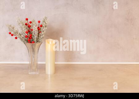 A beautiful arrangement features a clear vase with red berries and greenery beside a lit candle. The soft, neutral background enhances the warm ambian Stock Photo