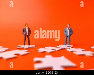 The image depicts miniature figurines of two businessmen standing on separate white jigsaw puzzle pieces against a bright orange background. Stock Photo