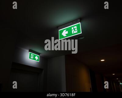 Green illuminated emergency exit signs in a dark hallway indicate escape routes for building safety. Useful for themes of evacuation, hotel security, Stock Photo